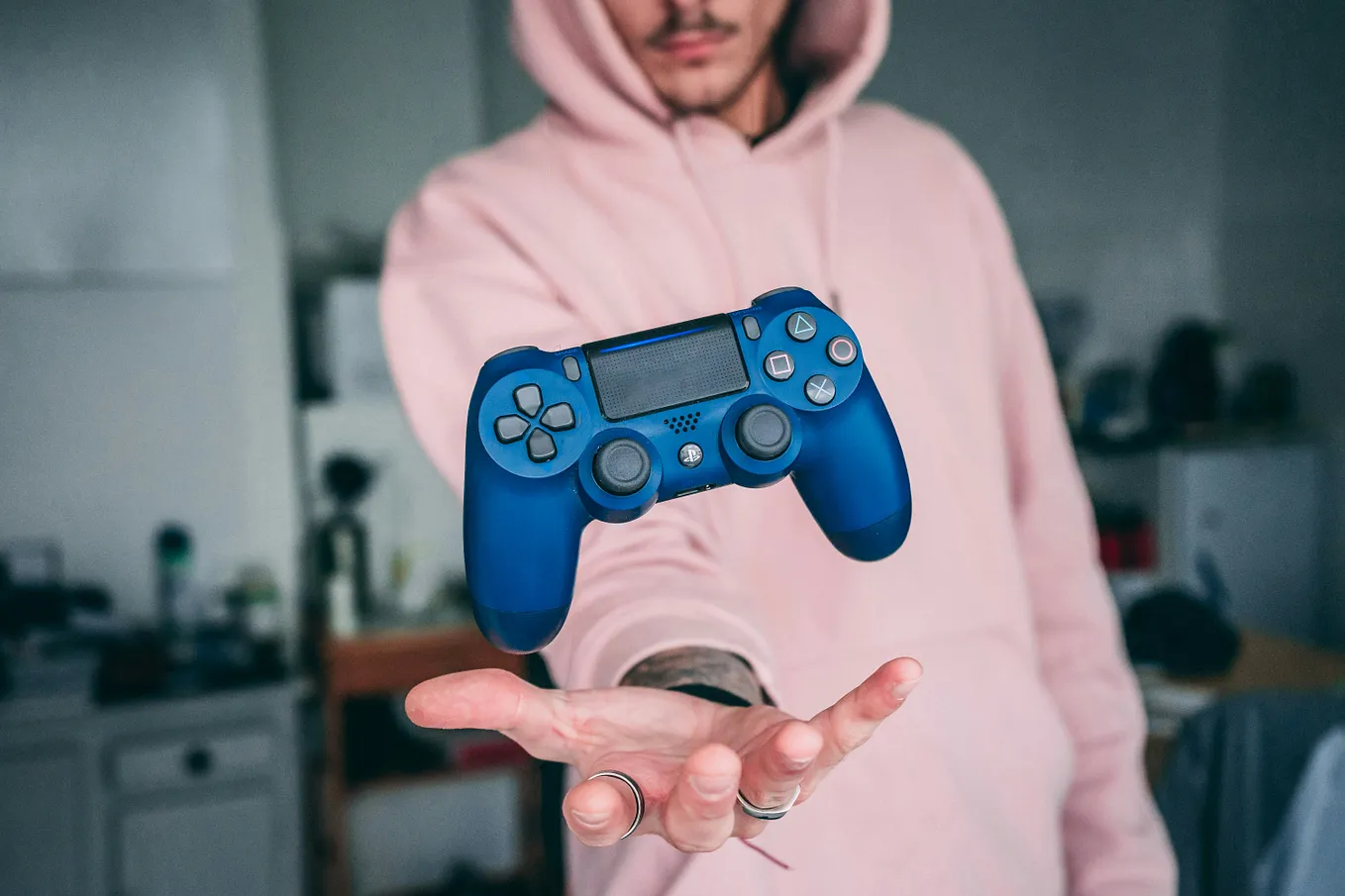 Image shows man’s hand holding blue video gaming console in focal point of photo. Background shows man with facial hair in pink hoodie.