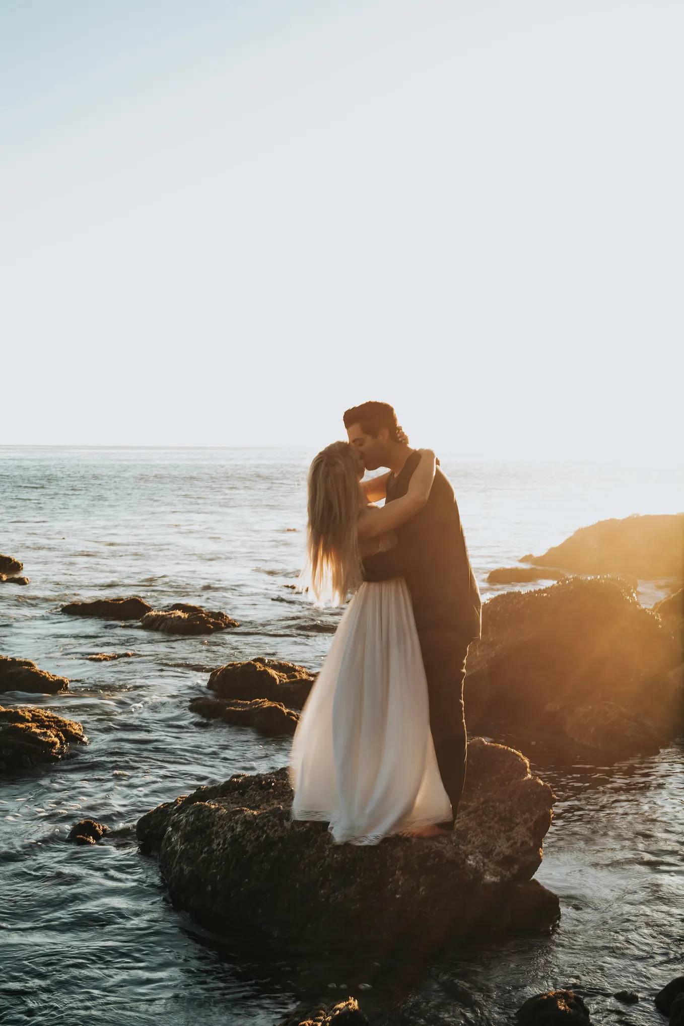 A man and woman kiss while standing on a rock in the ocean.