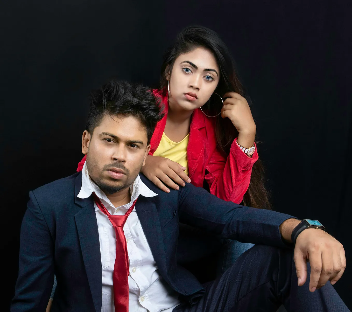 A man and a woman in office attire in power poses. The woman is wearing a red suit with a yellow hirt, and man is wearing a blue suit with white shirt and red tie.