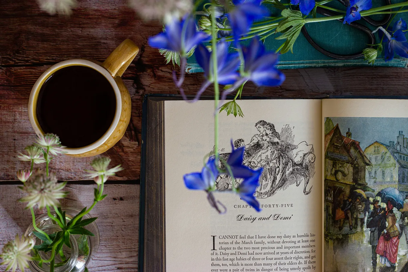 A bird’s eye of a table top with a coffee mug on the top-left quadrant and an open copy of Little Women by Louisa May Alcott on the right-side. Violets and white seedling blossoms frame the book.