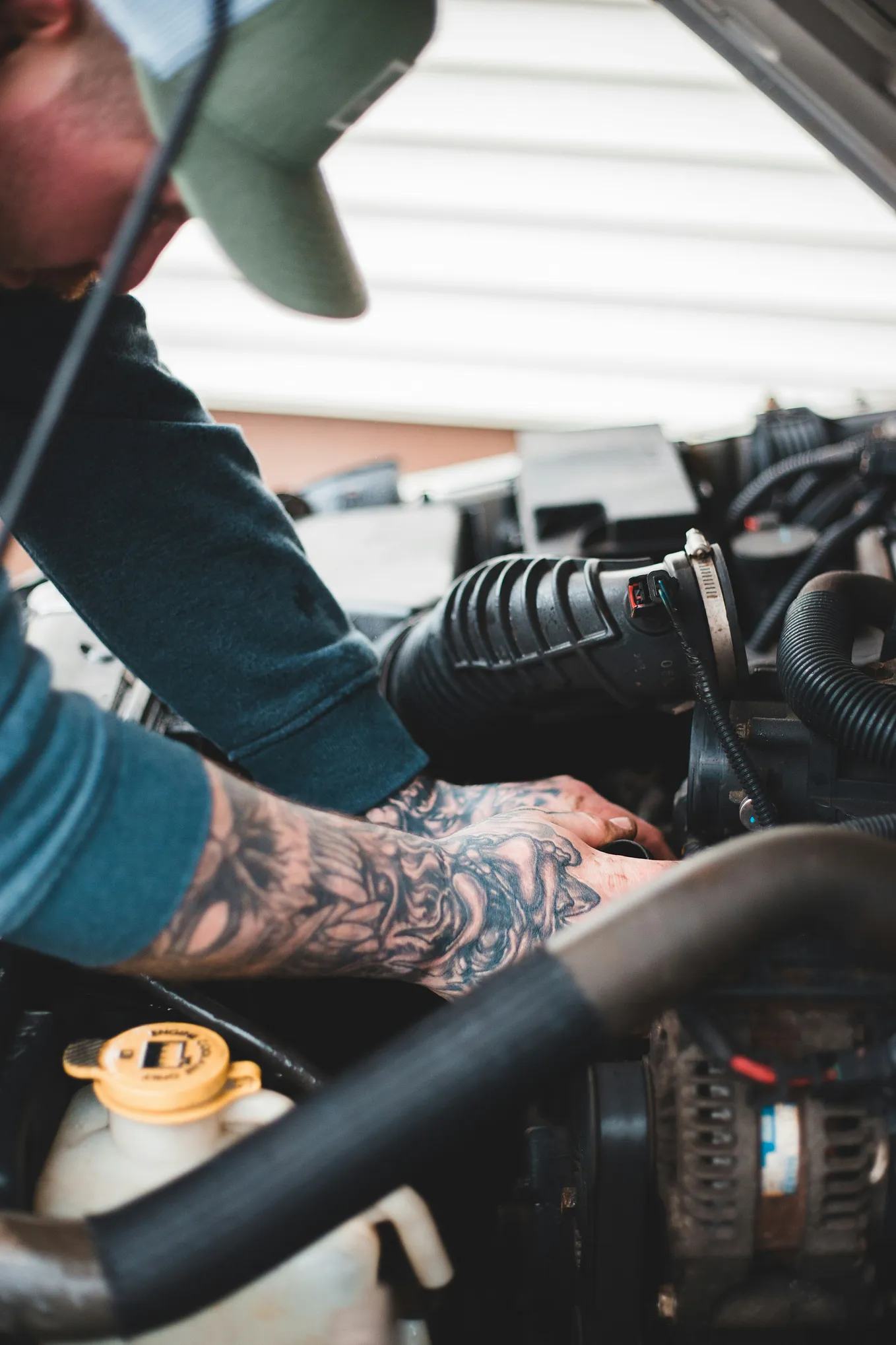 A man with heavily tattooed hands and wearing a baseball cap works at a car engine.