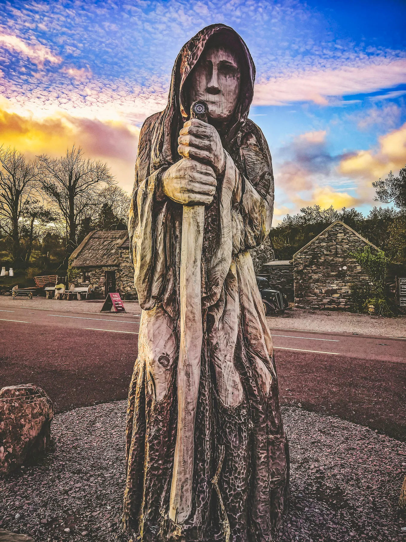 Carved wooden image of an old woman in a cape with a staff against a backdrop of winter trees, a stone building and sky dappled with pink and yellow clouds