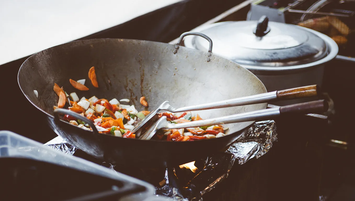 steel asian wok on a gas burner with pieces of meat coated in sauce inside of it and a metal spatula resting on the wok