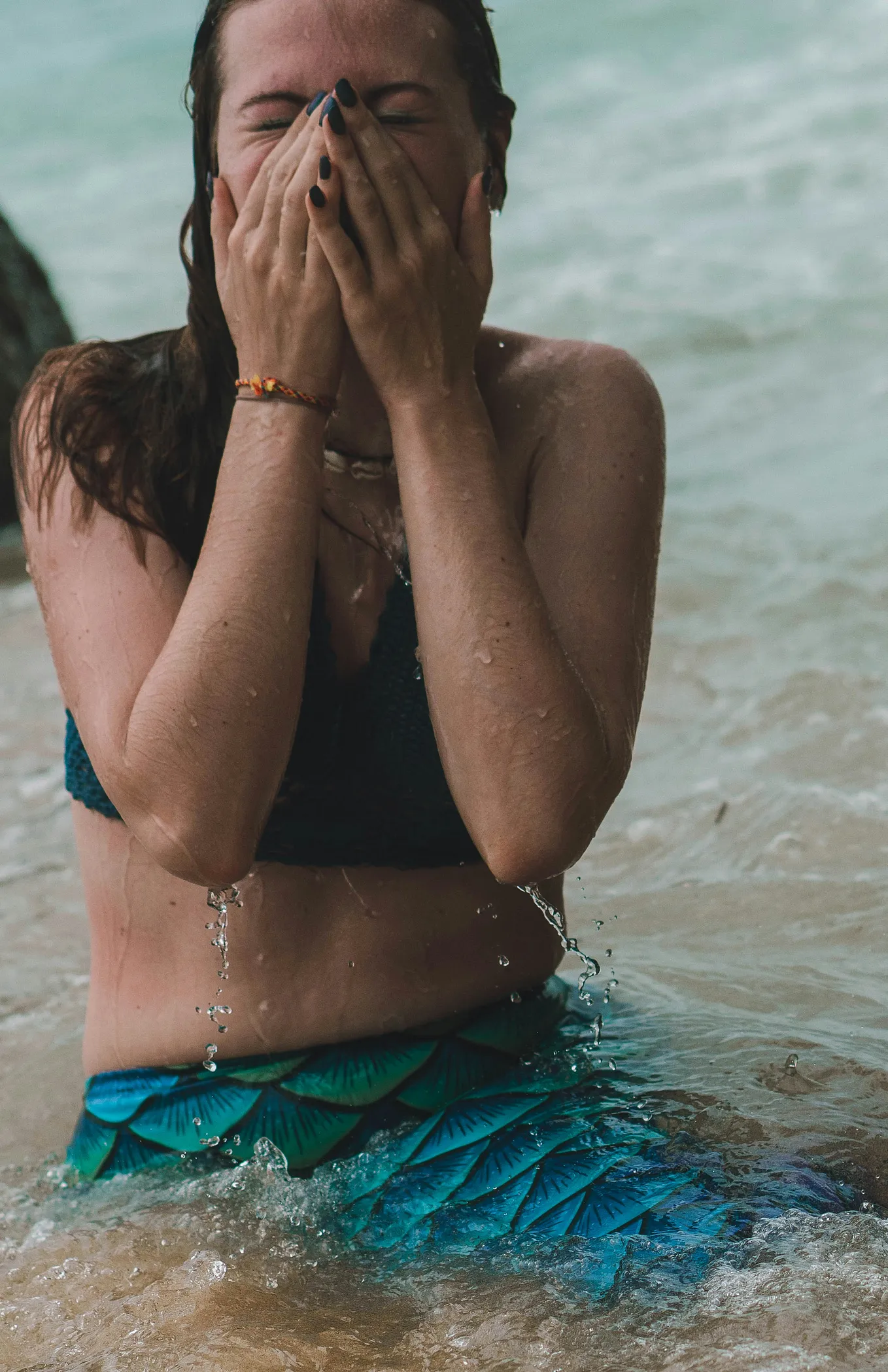 A woman dressed as a mermaid sits in the waves of the ocean and cries forcefully into her hands.