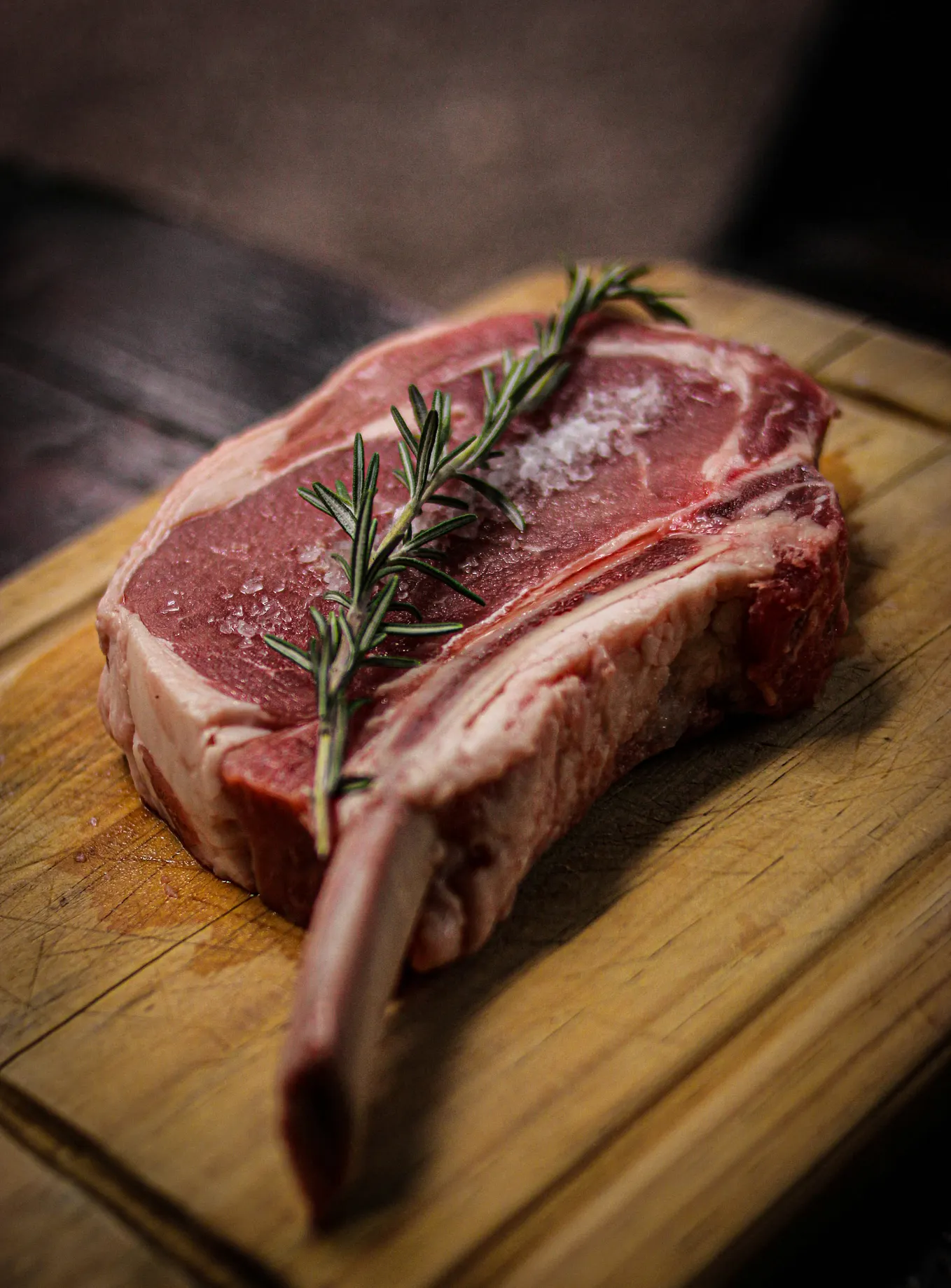 An extremely fatty, uncooked, bone-in ribeye, salted and sitting on a wooden board, topped with a rosemary sprig.