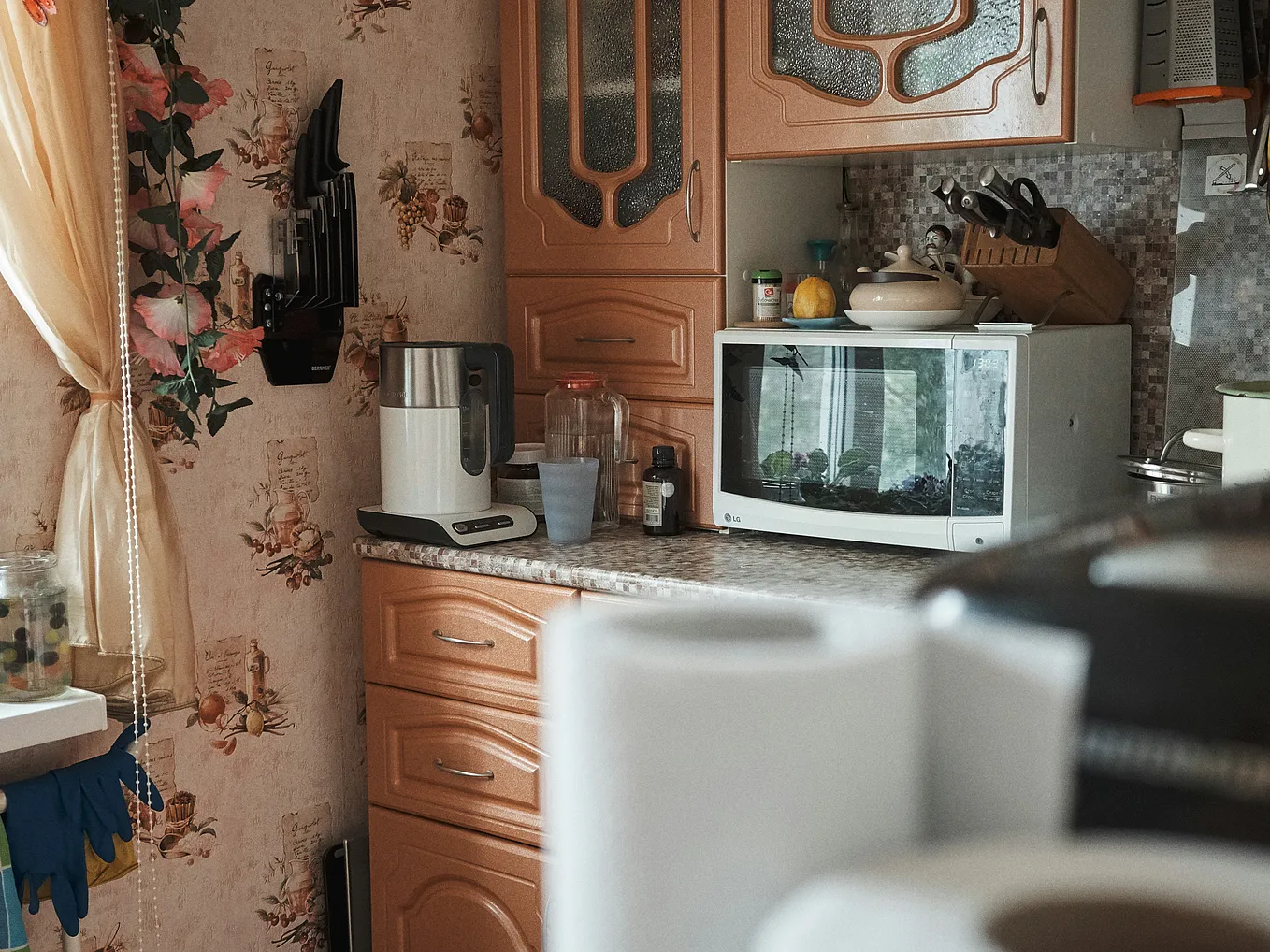 A cozy kitchen corner featuring wooden cabinets with ornate patterns, a floral wallpaper design, and a small counter holding various items. On the counter are a microwave, a knife set mounted on the wall, a glass jar, a kettle, and other kitchen tools. The window draped with light curtains lets in natural light, highlighting the warm and vintage aesthetic of the space.