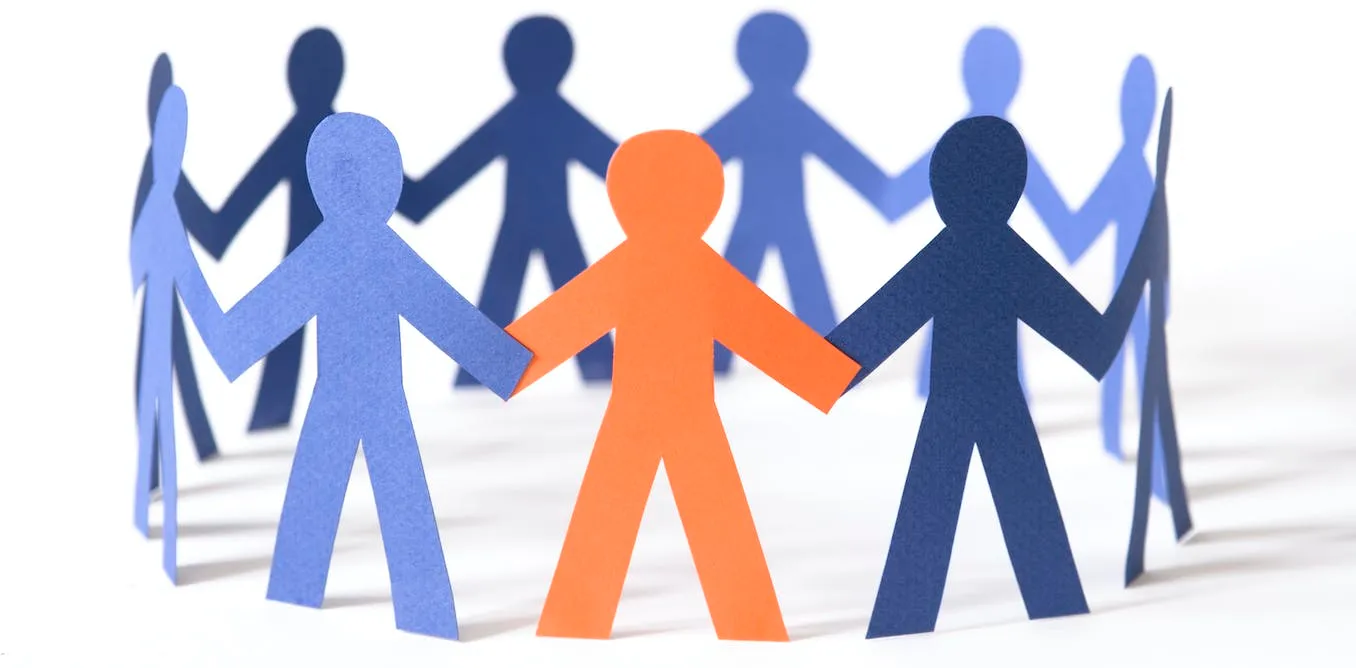 A circle of paper cut-out figures holding hands, one of them colored orange, the rest various shades of blue