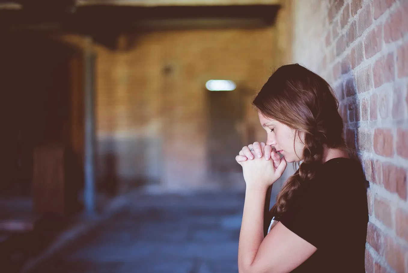 Young teenage girl with brown hair in a braid. Wearing a short sleeved black T-shirt, she is leaning against a brick wall, holding her hands in a prayer position in front of the lower part of her face. The eyes are closed.