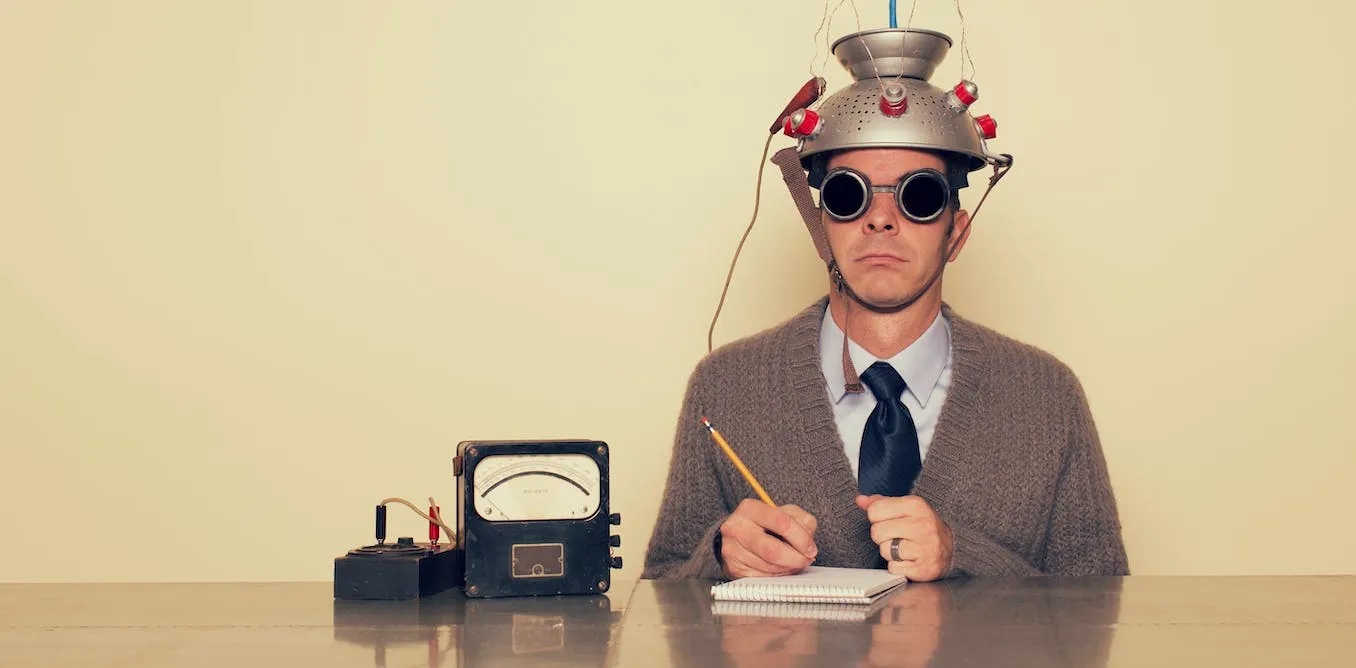 A person in a tin hat with lots of wires coming from it, reading a notebook