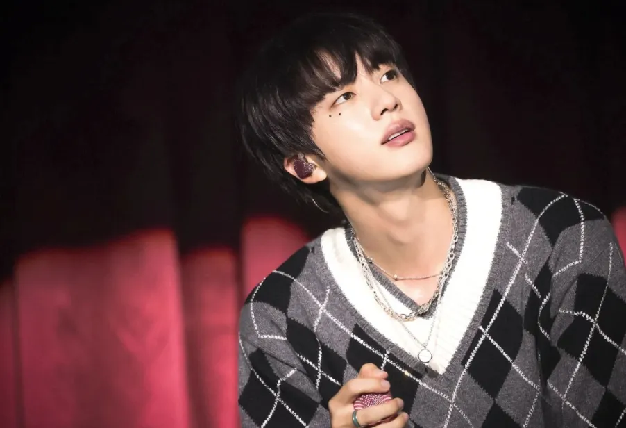 Jin of BTS on a stage with a red curtain behind him. He is looking up and he wears a grey and black argyle sweater with a white t-shirt undernearth. His face is decorated with a few pieces of glitter under his eye.