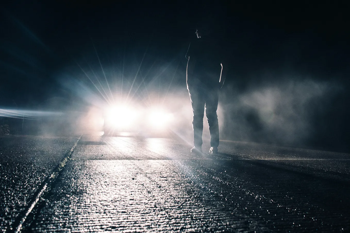 A man standing in front of a car headlights in the middle of the night.