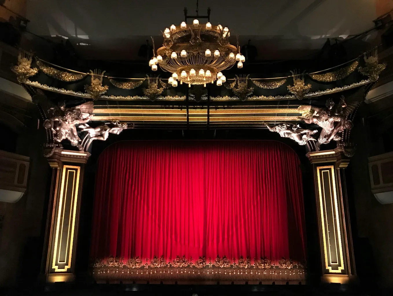 Theatre stage with red curtain, large candle chandelier hanging from the ceiling and gold framing around the stage