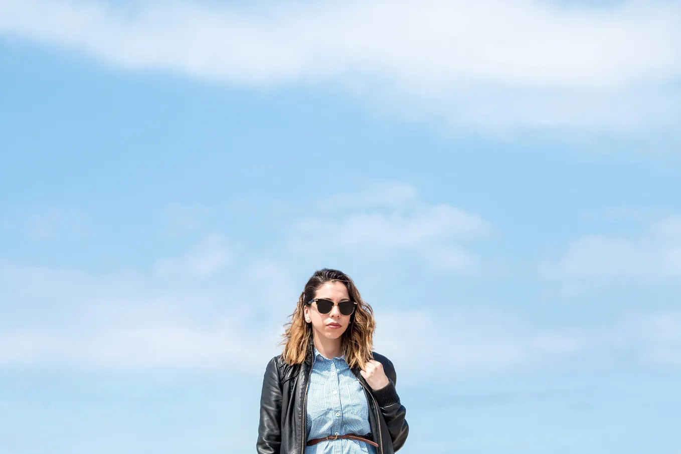 A woman in sunglasses and a leather jacket over a blue dress in the bottom of the frame with a beautiful blue sky behind her filling most of the frame