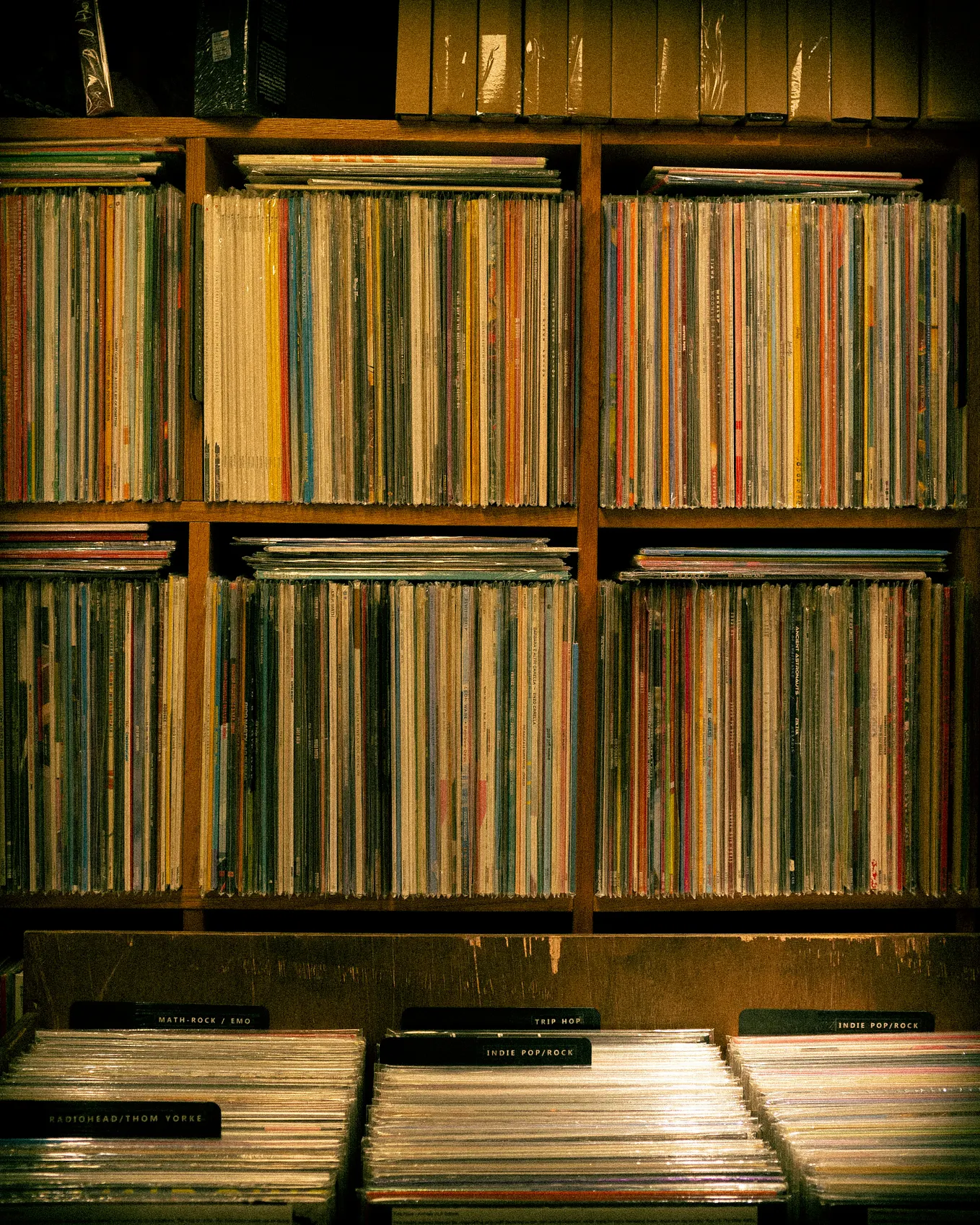 Wooden shelves packed with vinyl records organized by genre, warm amber lighting, with visible section labels including Math-Rock/Emo, Trip Hop, and Indie Pop/Rock.
