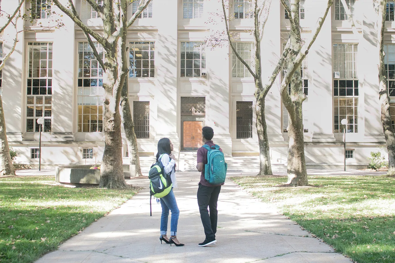A female and male student with rucksacks on their backs standing in front of a college building