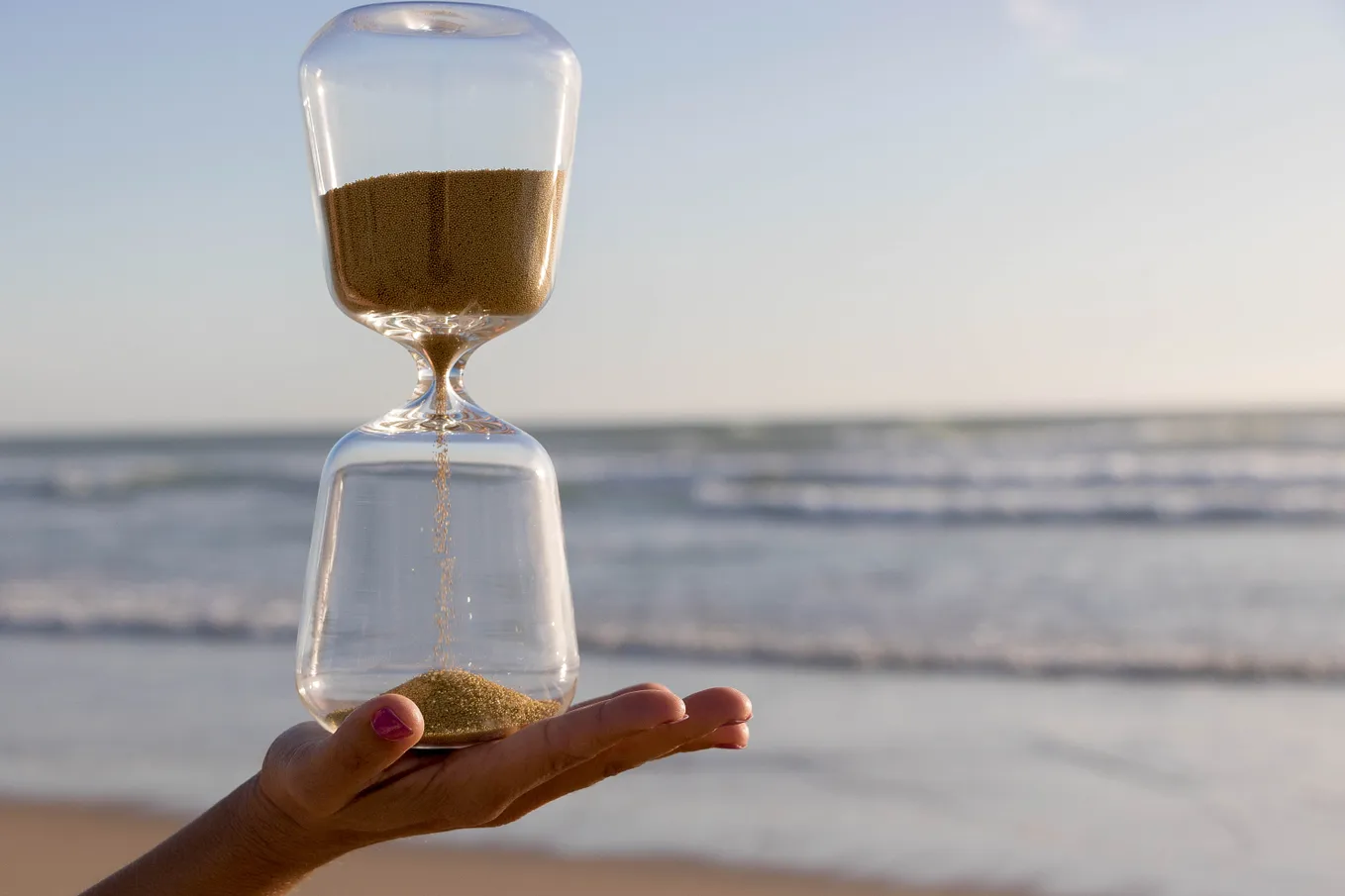 Hand holding an hourglass with sand running out; ocean and sky in background