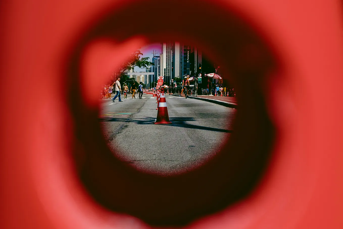 A street lined with traffic cones and filled with pedestrians as viewed through a hole in another traffic cone.