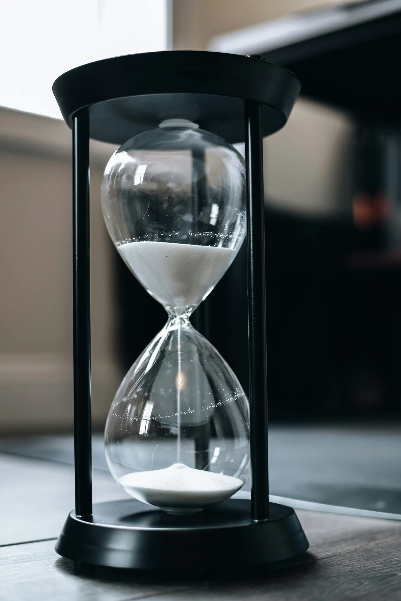 A black hourglass with white sand sits on a dark wooden table, mid pour