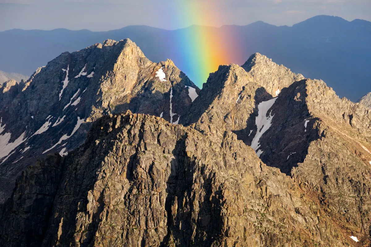 A rainbow soaring over a collection of rugged peaks with a blue mountain range in the background.