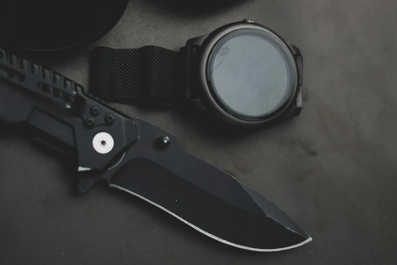 A black knife and a watch with a black band resting on a dark grey surface. The watch face is just a blur of white.