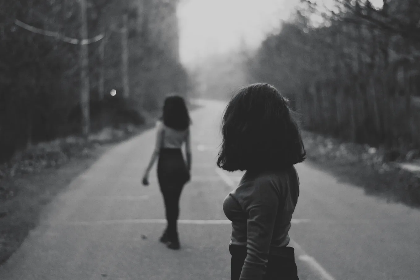 A young woman is looking back over her shoulder at another young woman who is walking away down an open road.