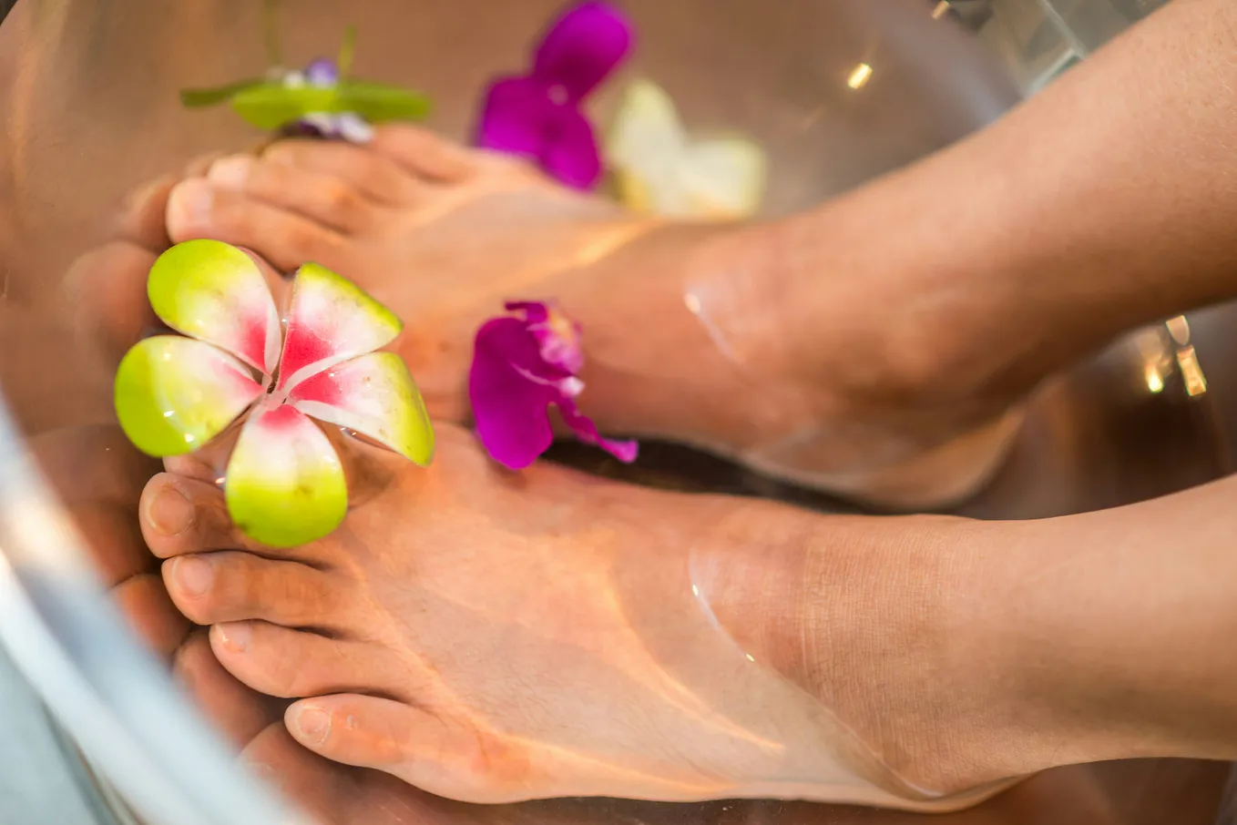 Light-skinned bare feet soak in a few inches of water. Pink flower petals float in the water, and one foot holds a white, pink, and green blossom between the big toe.