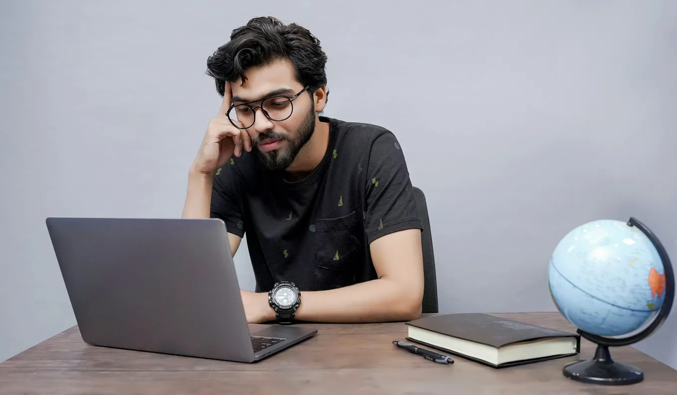 A student on a laptop, with a closed book, a pen, and a globe on their desk