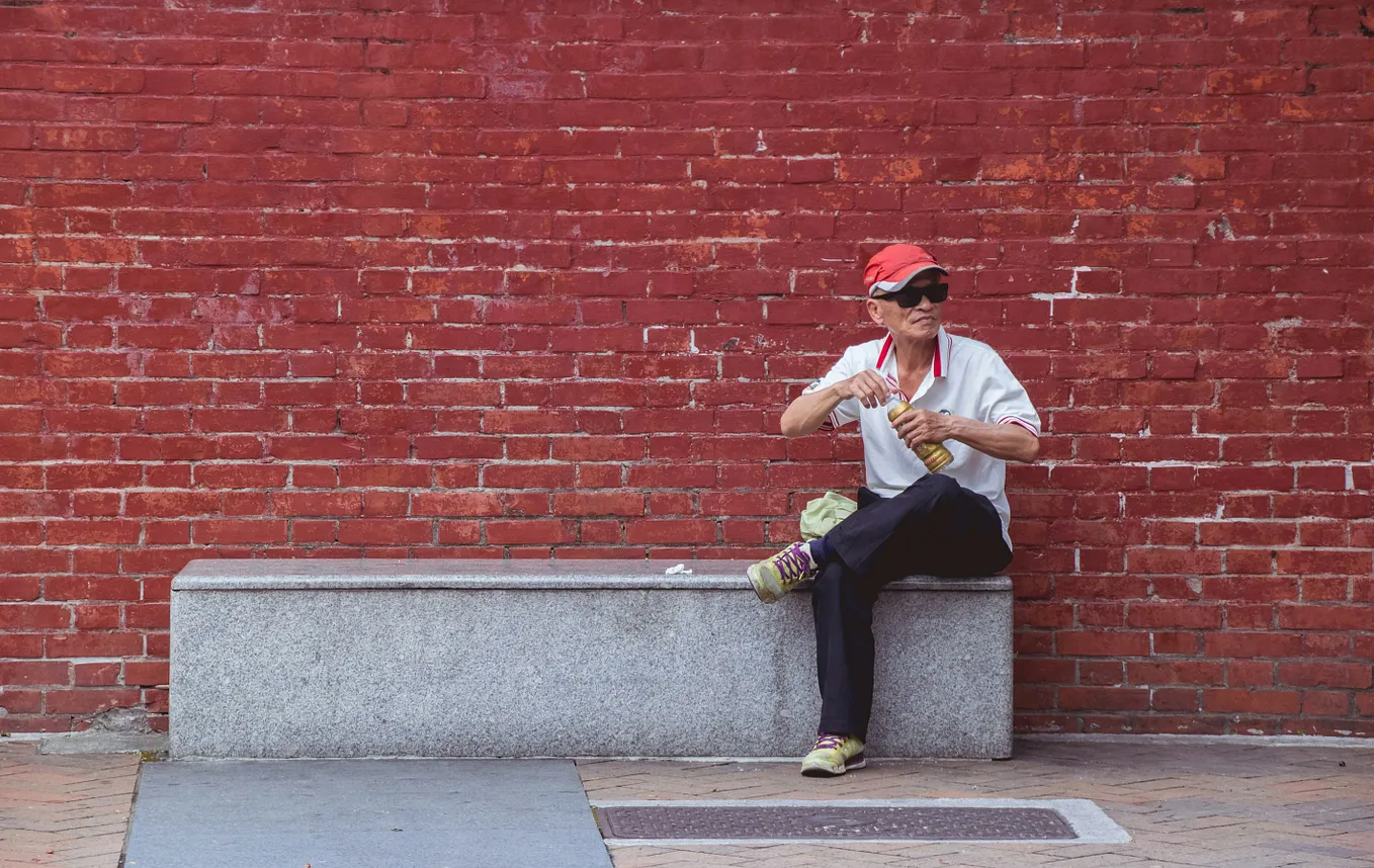 A man sits alone in front of bright red wall, opening a bottle.