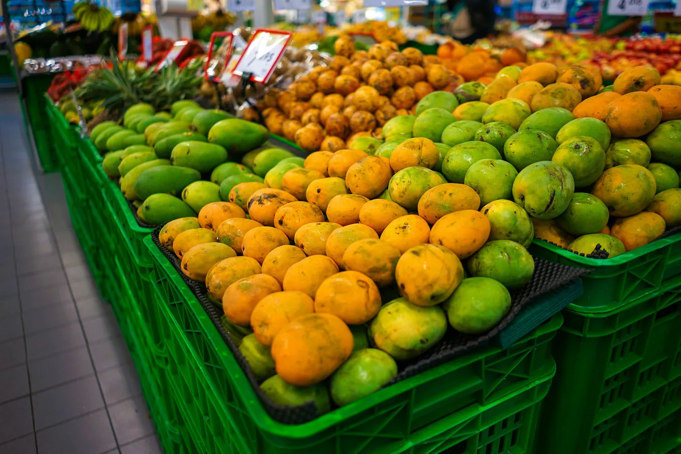 Coloured image showing the fruit section of a supermarket. Oranges and apples are displayed in green plastic containers.