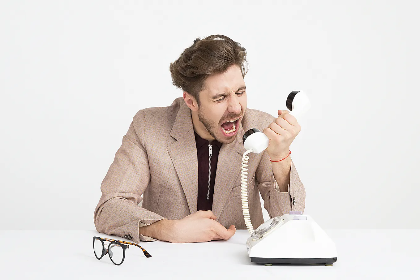 Angry white man in beige dress jacket with black shirt underneath. He’s holding a corded phone away from his head and appears to be shouting into it. A pair of glasses sit in front of him to the left and the phone base to which the cord is connected sits in front of him to the right. The phone is white with a black base and the ear/speaker piece he’s shouting into is white with black rim at the ear and mouth parts.
