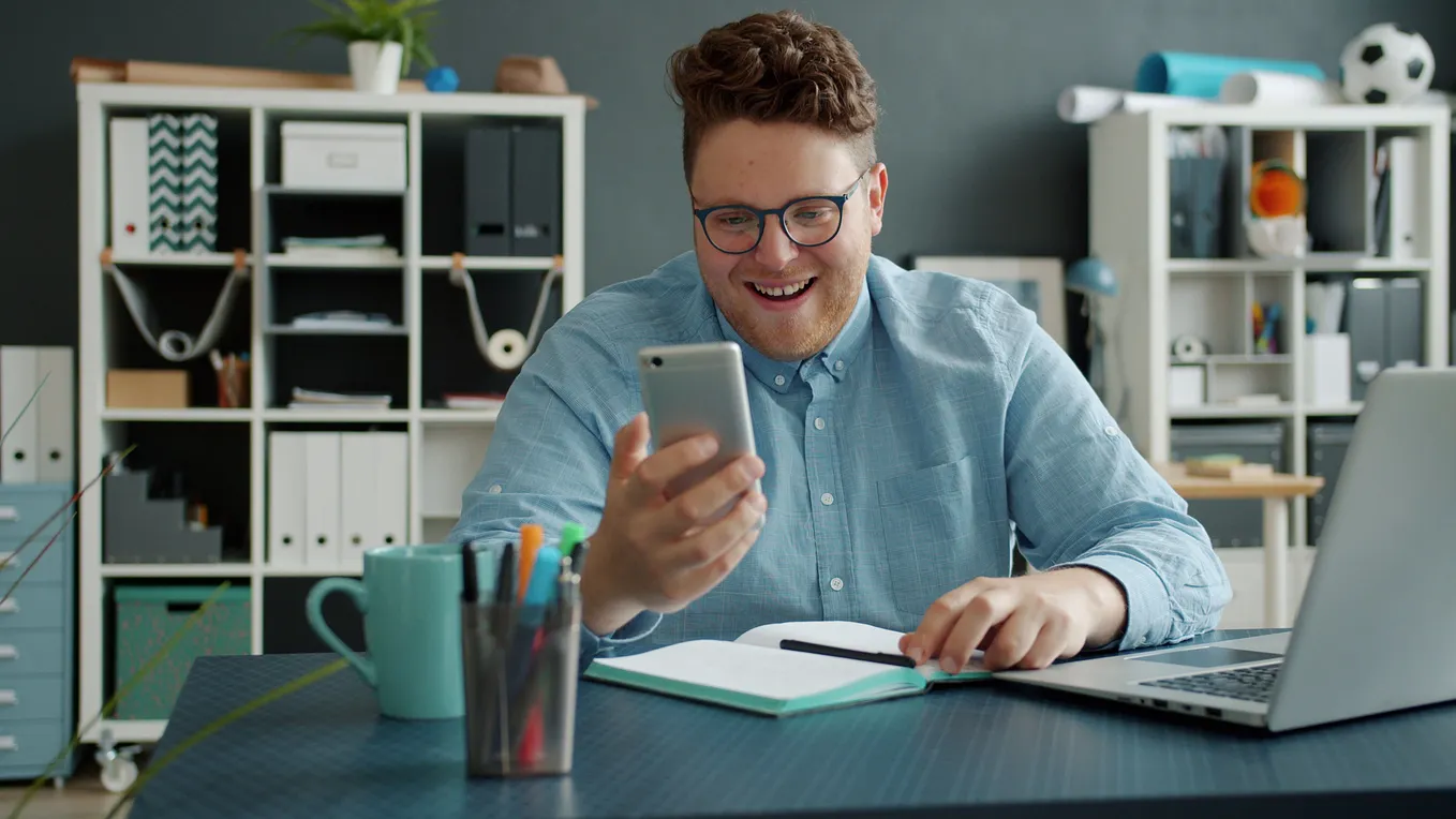 Cheerful man in glasses using smartphone at a desk with laptop.