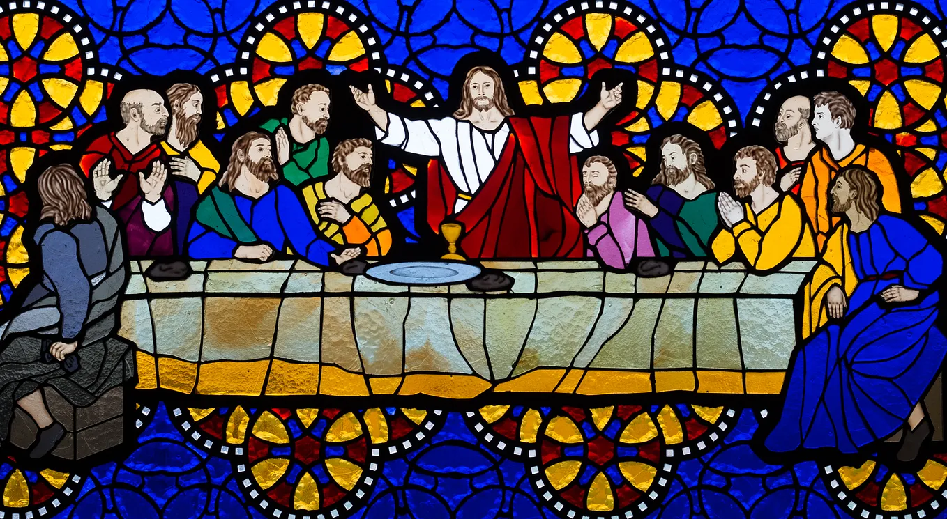 A colorful stained glass image of the Last Supper. Christ stands in a white robe with a red sash with his arms extended.