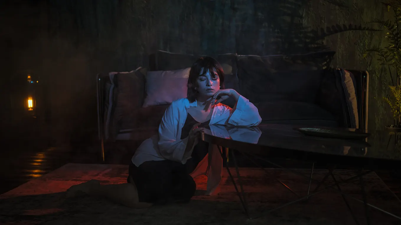 A worried woman sitting on the carpet in a dark sitting-room with a yellowish light bulb nearby and a sofa right behind her.