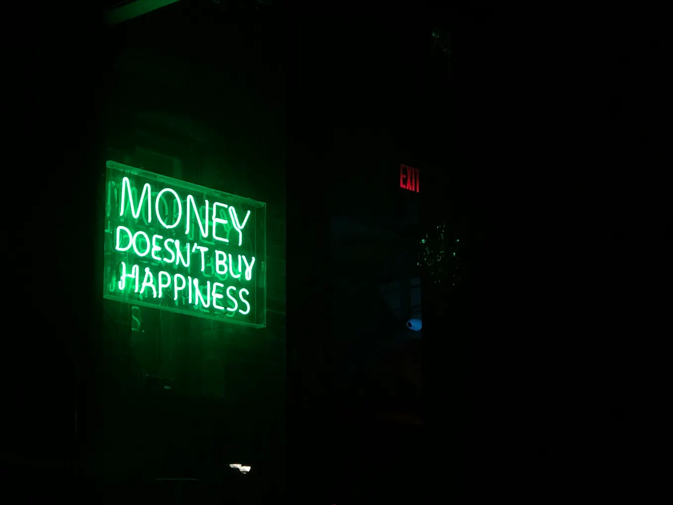 The image shows a green neon sign displayed in a dark room that reads “MONEY DOESN’T BUY HAPPINESS” in bright, glowing letters. The sign is mounted on what appears to be a wall, creating a striking contrast against the black background. To the right of the frame, there’s a red EXIT sign visible in the darkness. The atmosphere is moody and contemplative, with the neon green light casting a subtle glow on the surrounding area.