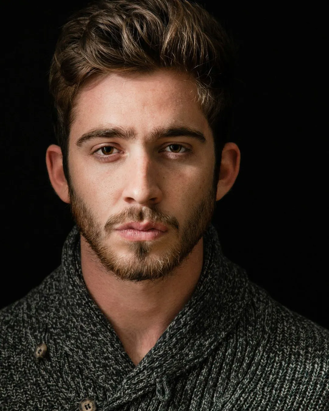 A non-plussed looking young man stares straight on. The portrait is a close-up. He wears a dark gray sweater and a beard of brown stubble.