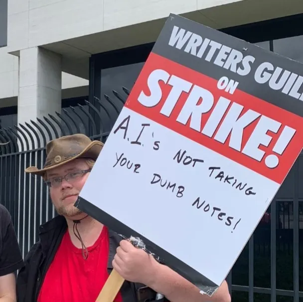 A picketer carries a sign reading “Writers Guild on Strike! A.I.’s not taking your dumb notes!”