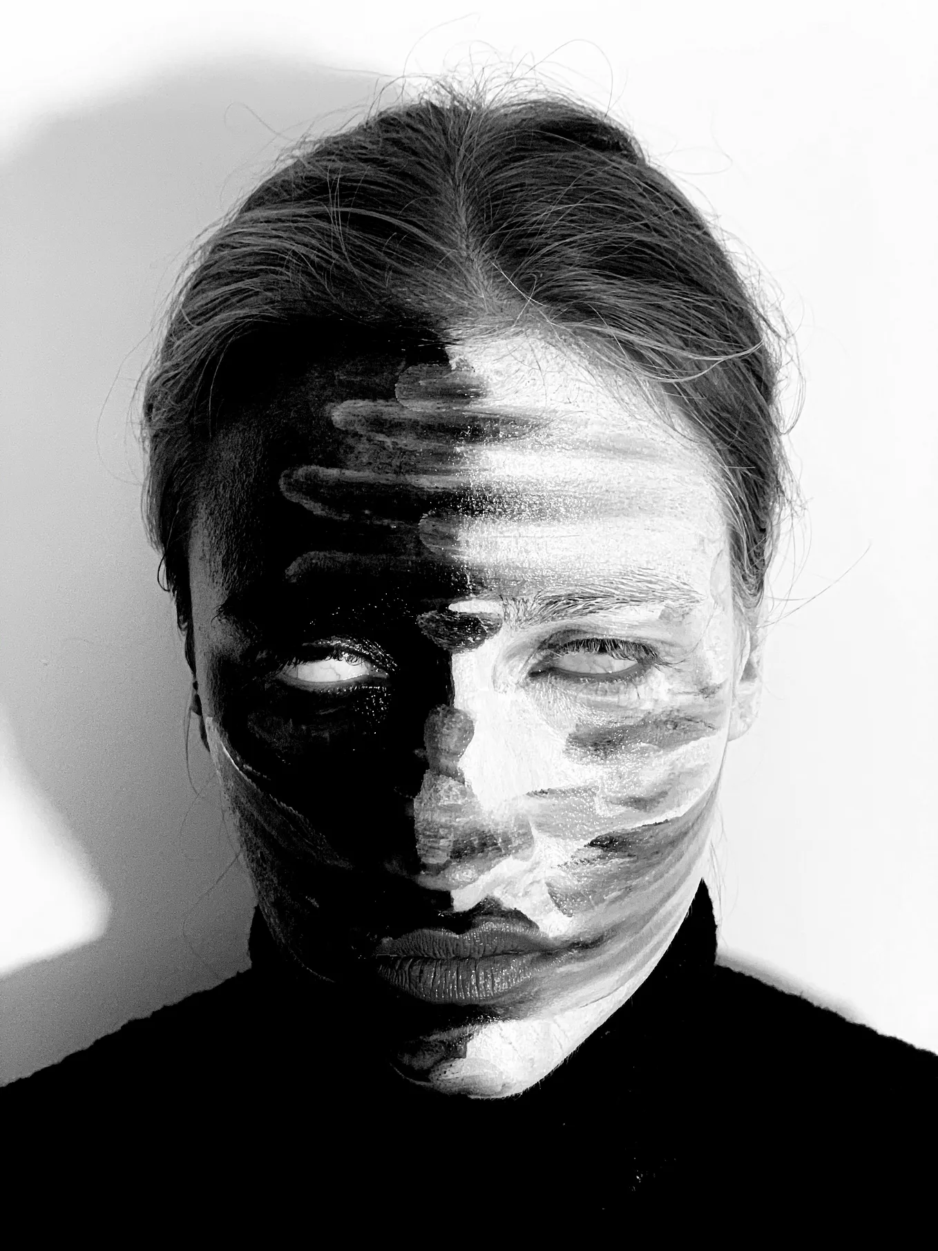 The image is a black and white portrait of a person with their face partially painted or cast in shadow, creating a stark contrast. The painting or shadow creates horizontal stripes across the face, which obscures the person’s features and gives a slightly abstract or artistic effect. The person is wearing a black top, and the background appears to be plain and light-colored, adding to the contrast.