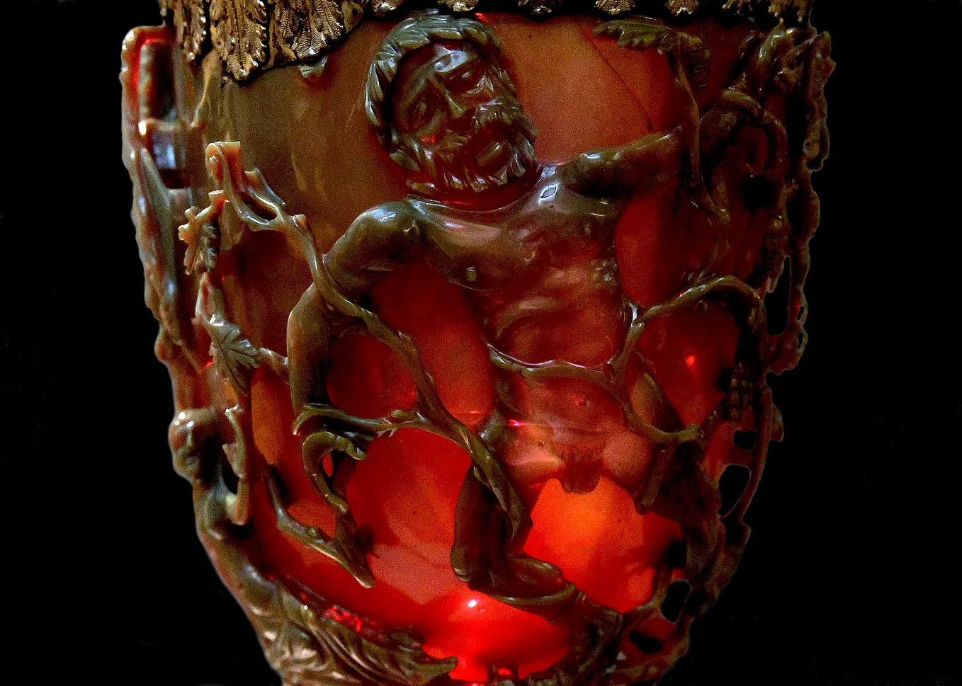 Engraved glass depicting a male bearded figure entangled in grape vines. The glass is jade green but has a bright red tint where the light shines through it, making it appear as it it is on fire from within
