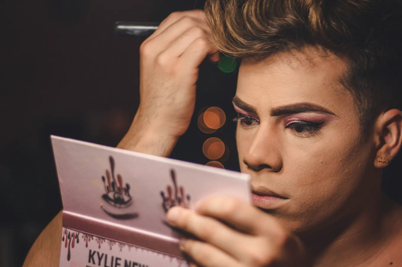 A performer applies makeup while using the mirror on their blush pad. Their eyebrows are slightly furrowed. They appear deep in concentration. They have colorful eyeshadow and winged eyeliner.