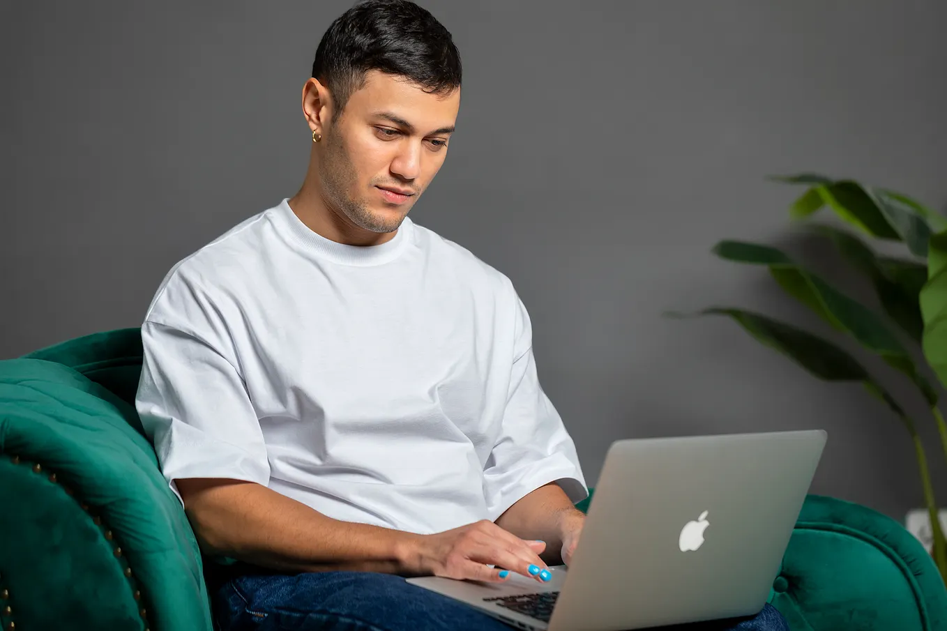 Photo of author working on laptap for work as a DEI consultant. They are Algerian and Amazigh. Brown skin, black hair, black eyes. They are seated on an emerald sofa, typing on their laptop which is sitting on their lap. They are wearing blue nail polish. The laptop is a grey Macbook air. The background is anthracite and there is a plant on the right side showing green leaves. The gaze of the consultant is down, towards the laptop.