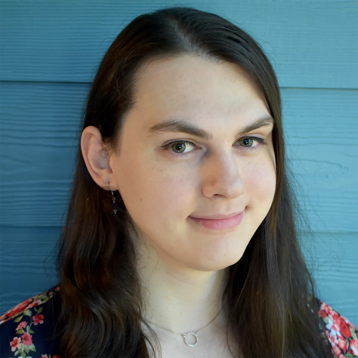 Picture of Tara Wilkins, a woman with long brown hair wearing a necklace and earrings, standing in front of a blue wall.