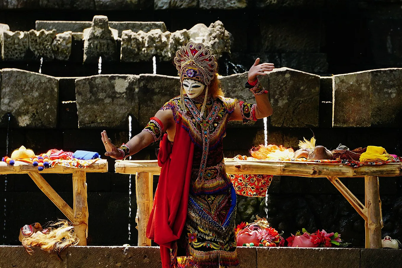 Colourfully dressed masked figure involved in an Indonesian ritual, in the background water streams through drilled holes in stone.