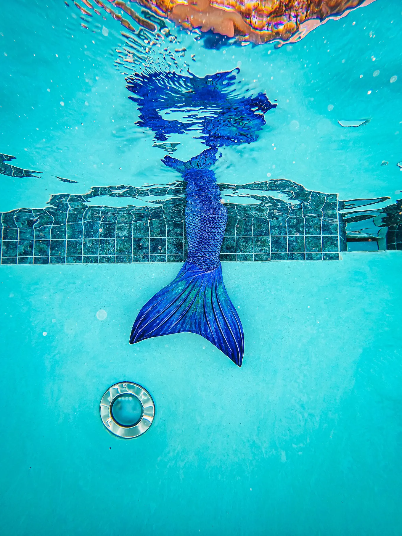 A blue mermaid tail dips from above, down into a swimming pool. Dark blue tiles are visible on the white pool wall, behind the tail.