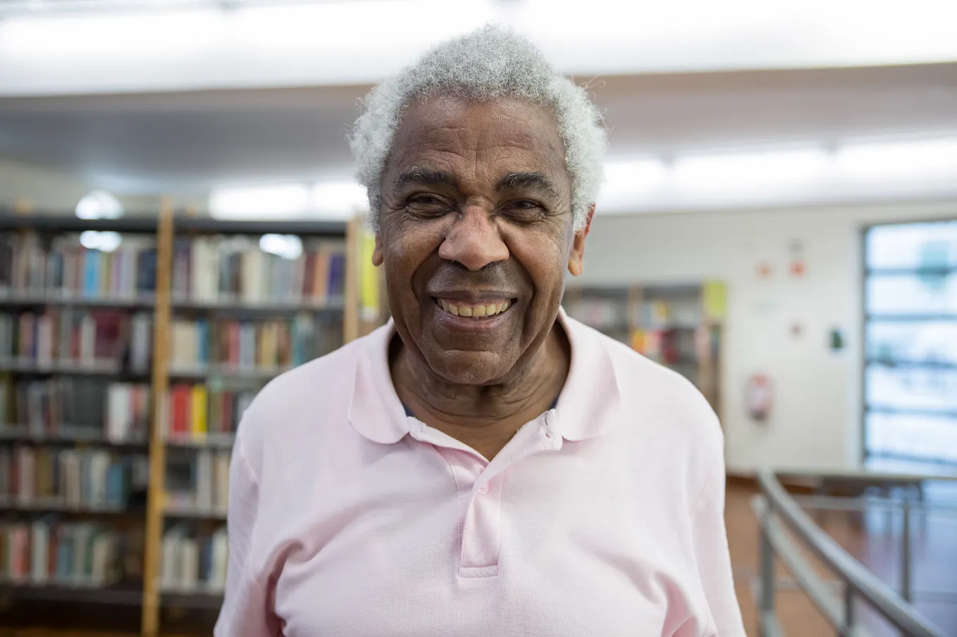 Elderly man smiling warmly inside a library. He’s wearing a pink t-shirt.