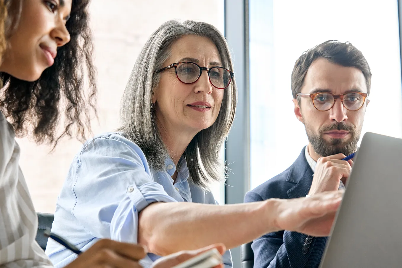 older woman in boardroom with a man and woman points at a laptop