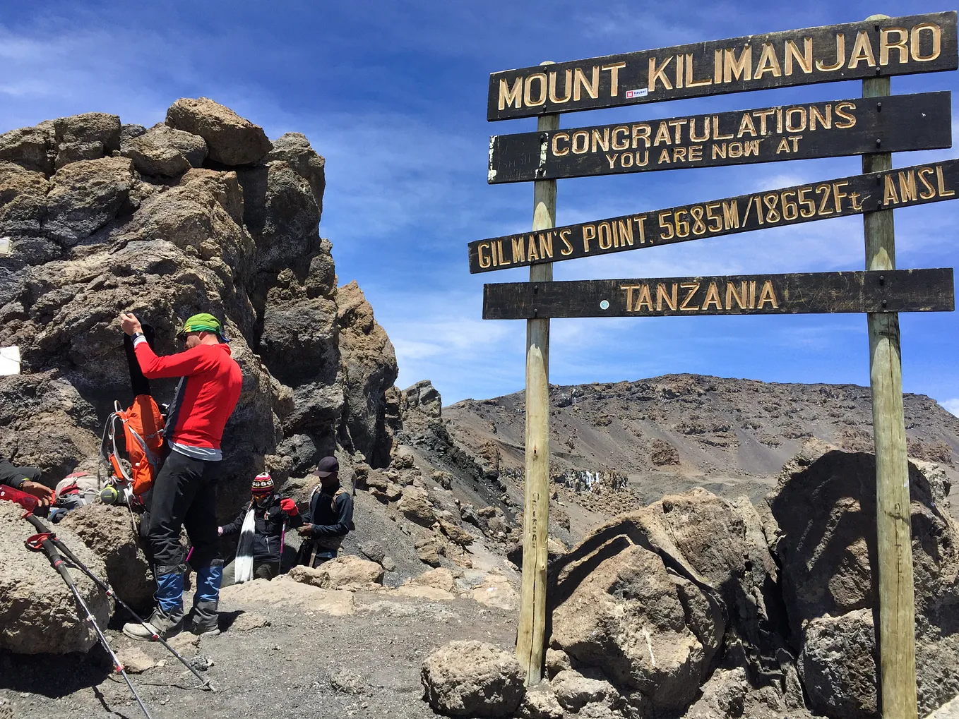Trekker stands next to a rock face putting items in his backpack. Signboards tell climbers they are now at Gilman’s Point on Mount Kilimanjaro