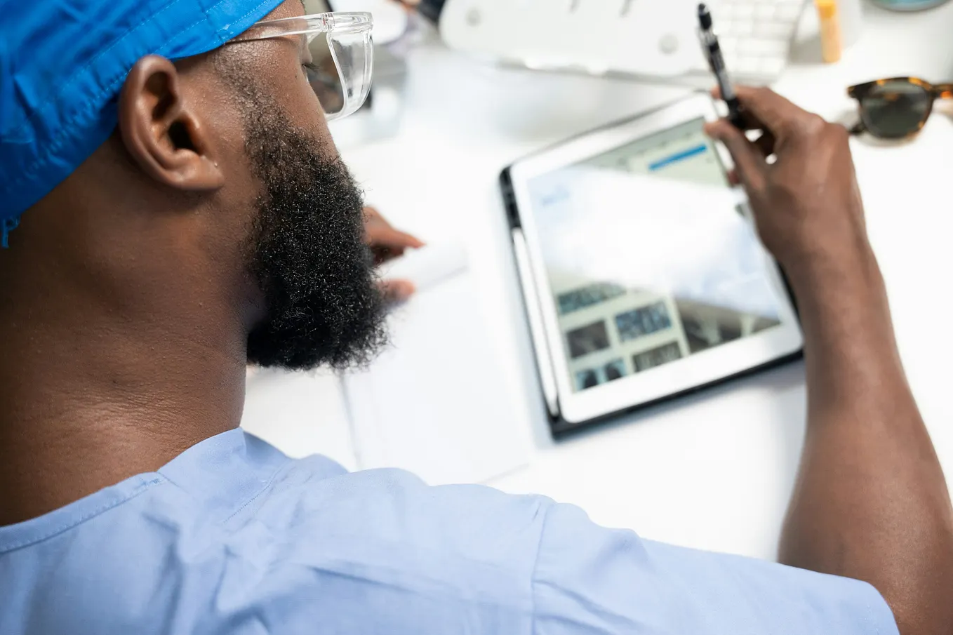 A man in a beard wearing medical scrubs while looking at an interface on a tablet and holding what appears to be a stylus