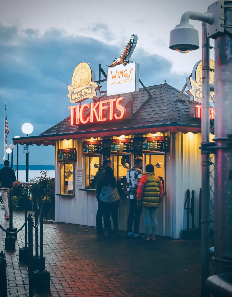 A ferris wheel ticket booth as seen on an overcast day with some patrons waiting in line to buy.