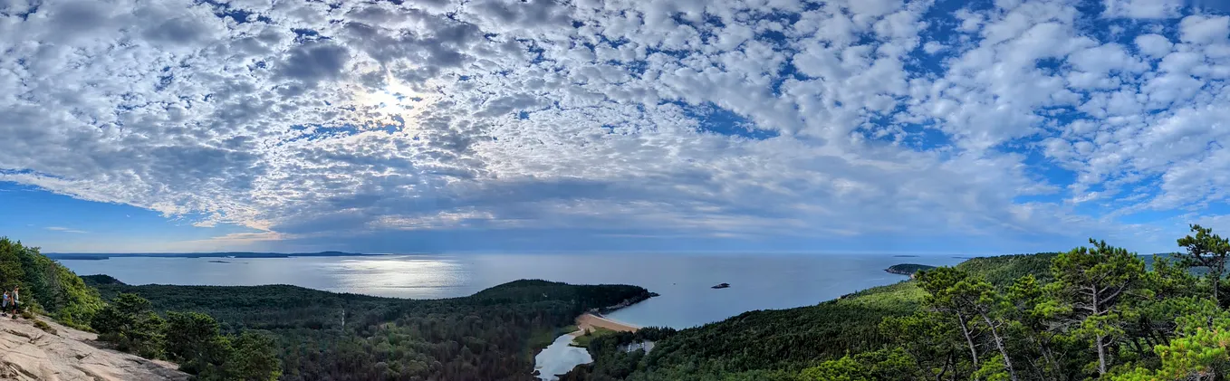 A sweeping scattered cloud cover takes up 2/3 of the top of the photo, and meets the blue ocean at the horizon. The bottom 1/3 of the image shows a rocky outcrop dropping to a green peninsula and mountain range with islands scattered in the distance