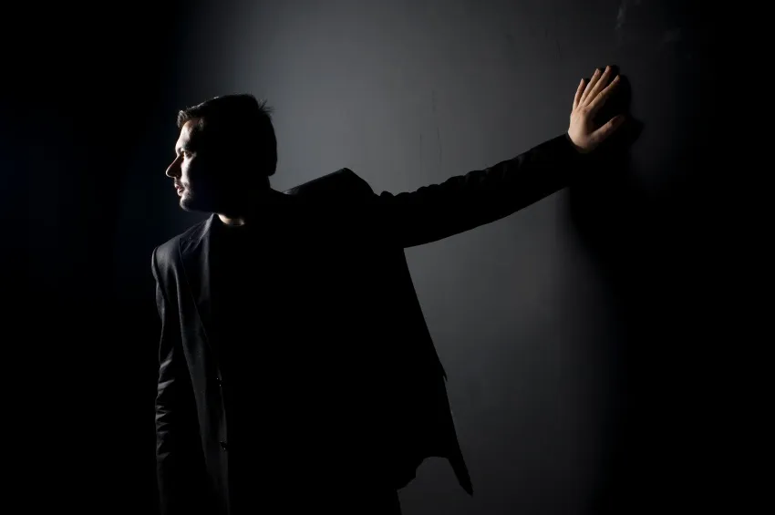 Image: A man in dark clothes and dim light, hand against the wall and looking the other way.